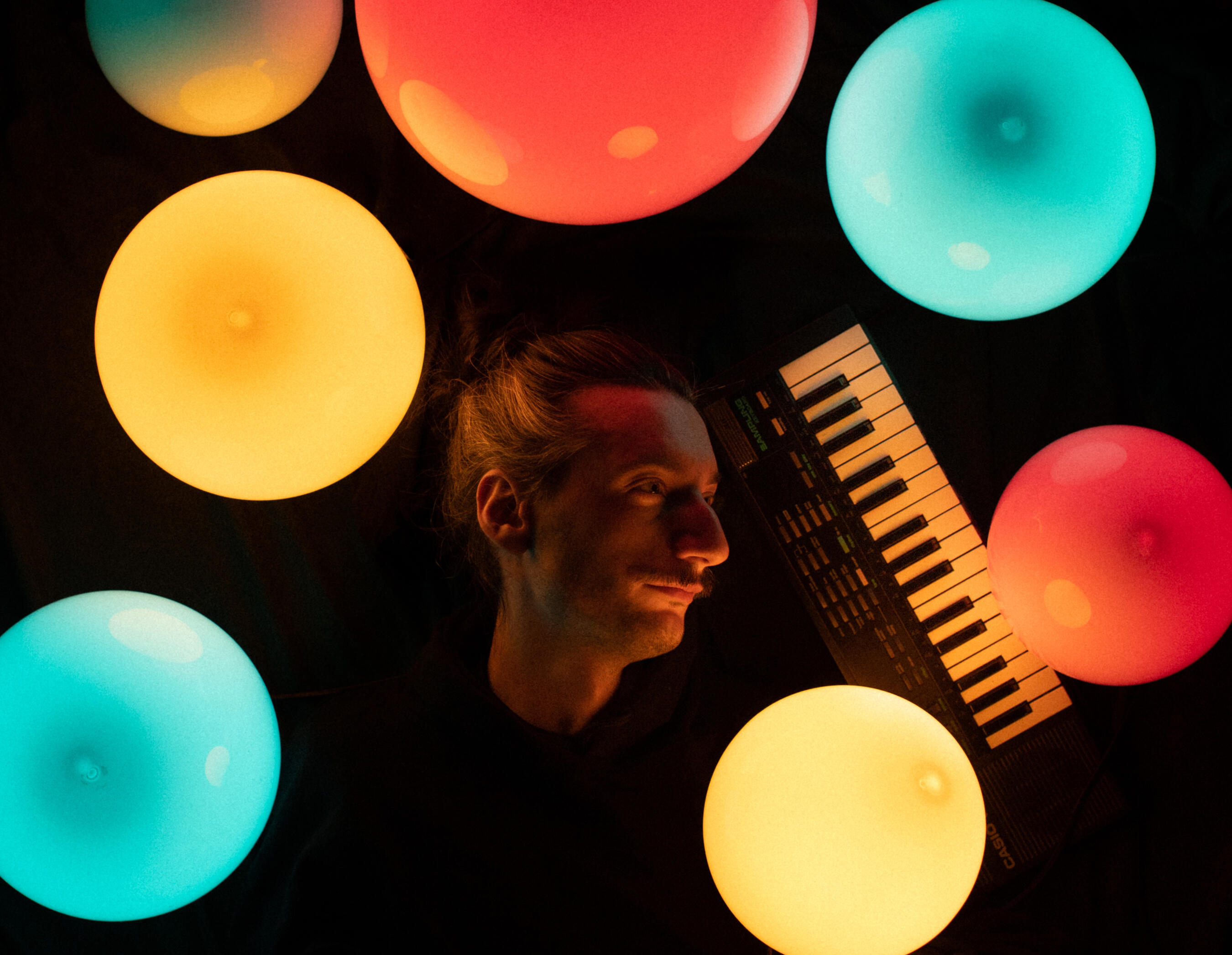 Overhead portrait of a musician lying in darkness beside a small keyboard, surrounded by glowing colorful orb lights in red, teal, and yellow.